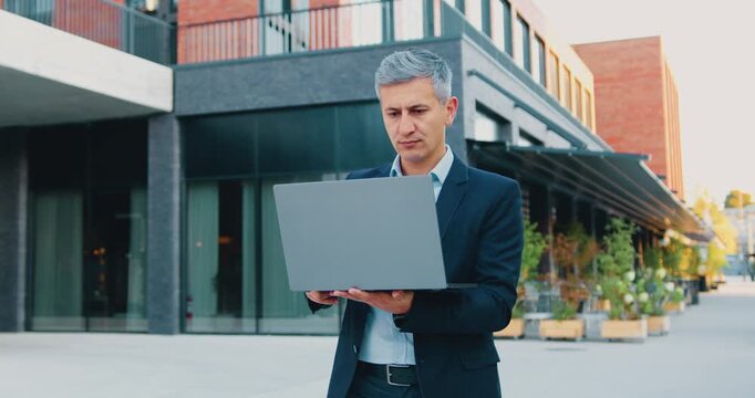 Businessman walking outdoors while working on laptop in front of modern office building, focused typing and subtle movement during daytime scene in urban courtyard, professional attire and serious