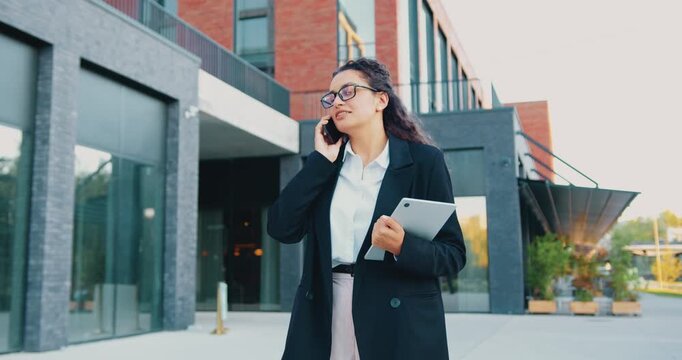 Confident woman professional standing outside a modern office building, holding a laptop in one hand and speaking on the phone with a colleague, engaged in a serious business conversation