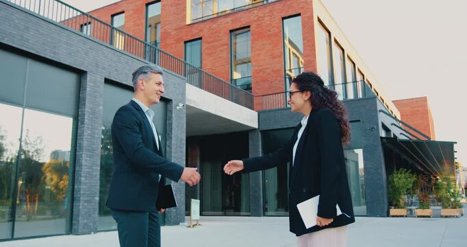 Close-up of two business partners, a man and a woman in formal suits, greeting each other with a handshake outdoors near an office building in the city.