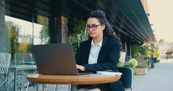 Young professional woman working on laptop at outdoor cafe table with focused expression and modern urban seating scene only visible in motion asset, daytime city terrace