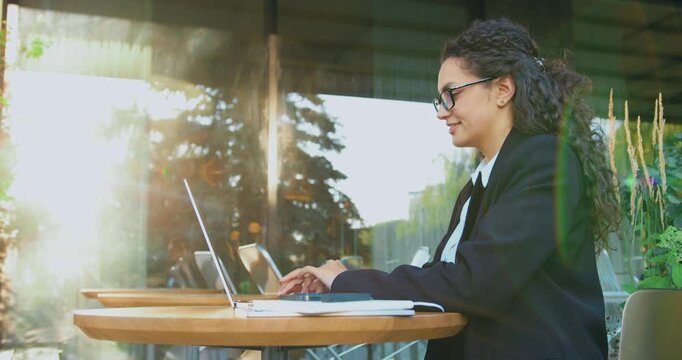 Smiling professional businesswoman typing on a laptop outdoors near a modern office building. Remote work concept, digital communication, corporate lifestyle, urban business environment.