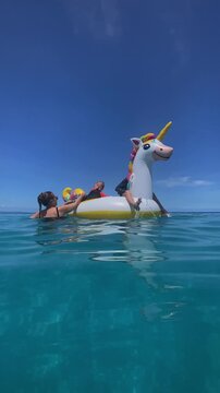 Children bounce joyfully. Children enjoying fun with colorful inflatable in ocean waves. Joyful kids bouncing on large unicorn float amid sparkling sea and bright sunshine, vertical video