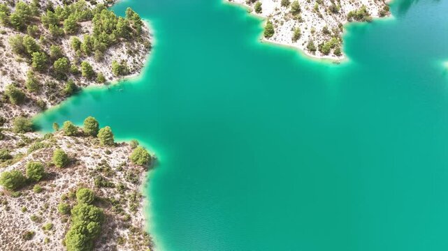 Aerial low over the blue green waters of Lake Portillo on a bright summer's day, following a channel surrounded by cream hillside dotted by native pine trees. Castril Granada Spain