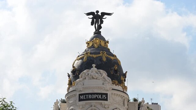 Telephoto view shows the famous Metropolis building dome in central Madrid, featuring rich gold ornate reliefs, classical marble sculptures, and the Winged Victory statue against a bright sky.
