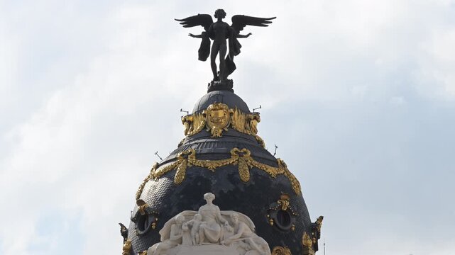Close-up telephoto shot highlights the bronze Winged Victory statue crowning Madrid's Beaux-Arts Metropolis dome, revealing gilded leaf garlands and marble allegorical sculptures in daylight.