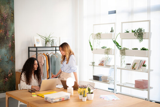 Two female colleagues collaborate in a bright, plant-filled modern studio. One woman types on a laptop while the other stands nearby, reviewing documents and discussing a project.