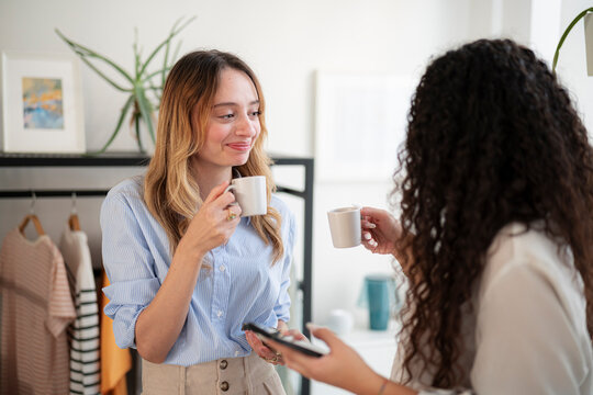 Two young women stand together in a modern apartment, drinking coffee from small white mugs and talking while holding a smartphone. They enjoy a morning break.