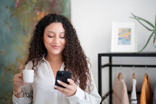 A young woman with curly brown hair holds a smartphone and a white coffee cup while smiling in a bright room. She reads a message on her phone during a relaxing morning.