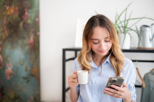 A young woman with blonde hair holds a white coffee cup while checking her smartphone inside a bright, modern room. She smiles pleasantly at the mobile screen.