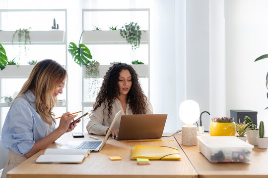 Two female colleagues work together in a bright modern office. One woman types on her laptop while the other holds a phone and takes notes at their wooden desk.