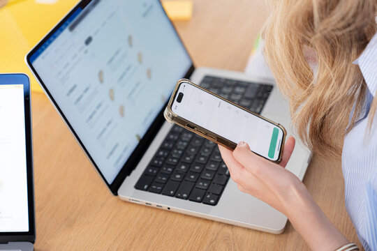 A blonde woman sits at a wooden desk and uses her smartphone while working on a laptop. She orders food online through a delivery app during a break at the office.