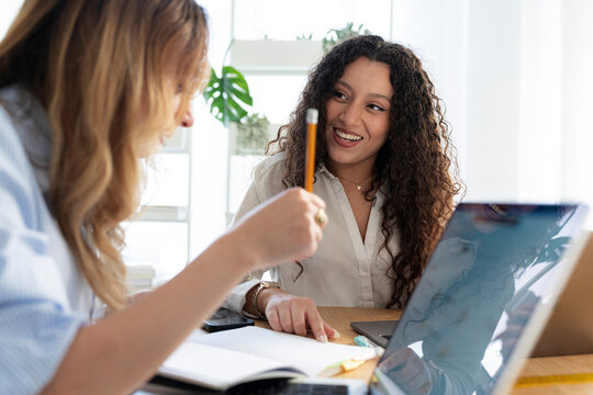 Two smiling female coworkers collaborate on a project in a bright office. One woman holds a pencil, pointing at an open notebook while discussing work with her colleague.