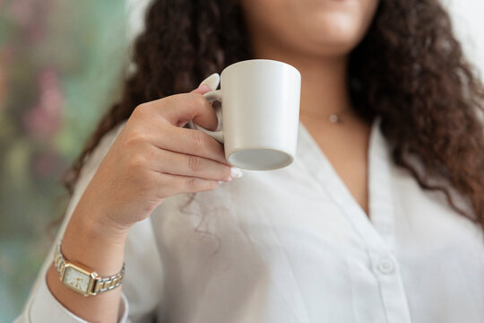 A young woman with long curly hair wearing a white shirt and watch holds a blank white coffee mug. She takes a relaxing morning break indoors, gripping the cup in her hand.