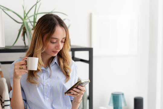 A young woman in a blue striped shirt holds a white mug while looking down at her smartphone. She is relaxing at home in a bright living room, checking messages and drinking.