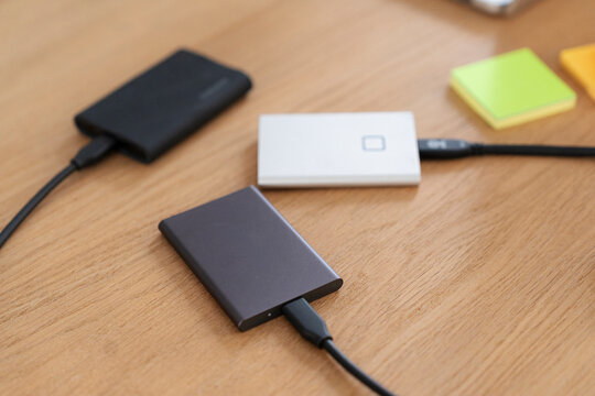 Three external portable hard drives are connected with cables on a wooden office desk. A person uses these data storage devices for digital backups and file transfer work.