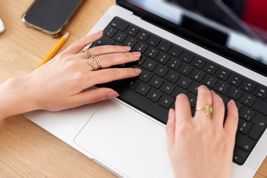 A woman wearing stylish rings types on a laptop keyboard at a wooden desk. Her hands rest on the keys while working remotely. A smartphone and pencil lie nearby.