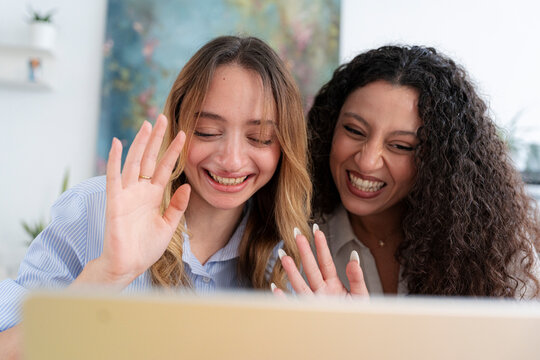 Two cheerful young women are sitting together at a bright desk. They smile broadly and wave their hands happily while talking on a video call using a laptop computer.