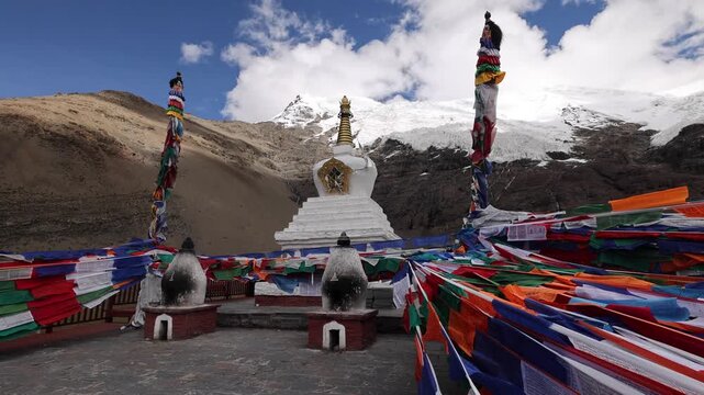View of the white stupa adorned with colorful prayer flags against the backdrop of the snow-capped Karo-la Glacier in Lhasa, Tibet, China.