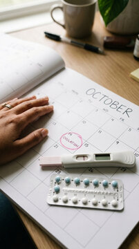Womans hand on calendar marking ovulation pregnancy test birth control pills on desk