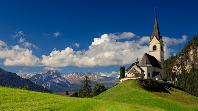 Schmitten, Switzerland - 21.09.2024: Traditional white alpine church with dark spire on green hill against snow-capped mountains and blue sky