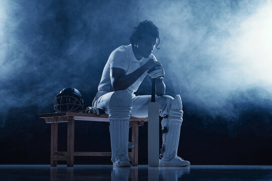 Thoughtful Indian batsman resting on bench beside helmet in smoky blue studio. Sports recovery, mental preparation concept.