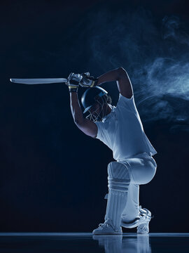 Committed helmeted batter following through on bent knee in smoky blue studio. Sporting discipline, batting performance concept.