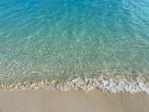 Aerial view of crystal clear turquoise seawater washing onto a pale sandy beach, revealing underwater sand patterns and gentle tropical waves.