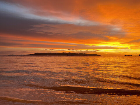 Vibrant orange sunset over the sea with dramatic clouds, gentle ocean waves, and a distant island silhouette creating a peaceful tropical coastal landscape.