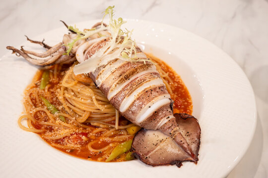 Grilled whole squid spaghetti pasta with tomato sauce on white plate.