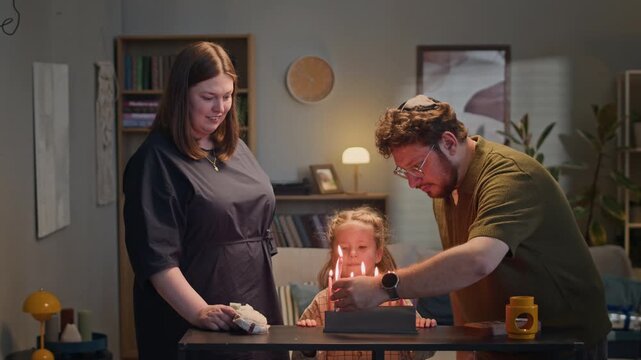 Caucasian parents and daughter lighting Hanukkah menorah candles at home and clapping. Clip suitable for Jewish holiday campaigns, cultural heritage marketing, and religious family storytelling