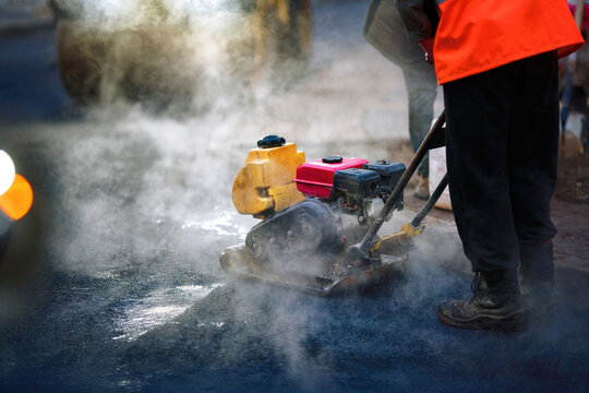 Night shift road construction worker operating plate compactor machine on hot steaming asphalt. Road repair process, rising steam as construction worker operates plate compactor during dark night work