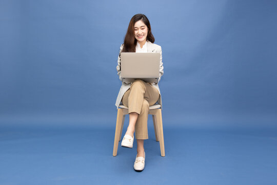 Smiling Asian businesswoman using laptop computer while sitting on chair isolated on blue background, representing online work, digital marketing, corporate connectivity, and modern remote job
