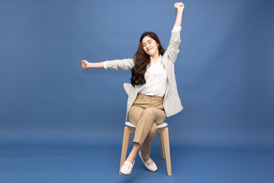 Cheerful Asian businesswoman stretching her arms while sitting on a chair against a blue background, taking a relaxing break to relieve stress and fatigue during work, promoting workplace wellness