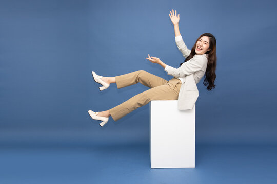 Excited Asian businesswoman sitting on a white box against a blue background, raising hands with joyful pose to celebrate career success, business achievement, promotion, and marketing proposal