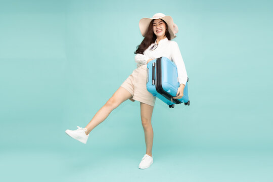 Cheerful Asian female tourist wearing hat holding a blue luggage suitcase isolated on mint green background, standing with excited dynamic pose for summer vacation journey, flight, and travel concept
