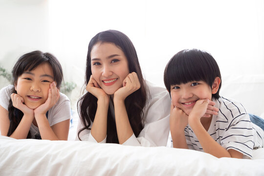 Happy Asian mother lying on bed with her son and daughter, smiling at camera with chin on hands to celebrate Mother's Day, family togetherness, love, childhood, and warm home relationship