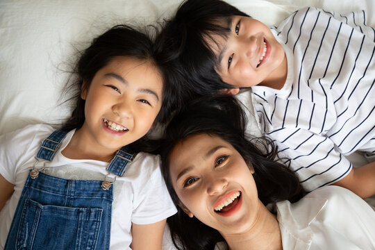 Top view portrait of happy Asian mother, son, and daughter lying together on bed laughing with head to head, expressing family love, togetherness, childhood joy, and Mother's Day celebration