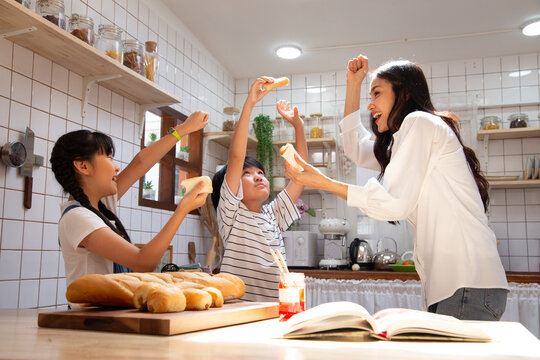 Joyful Asian mother playing and eating bread with her son and daughter in modern kitchen, raising hands with excitement for healthy family breakfast, playful morning meal, and warm home lifestyle