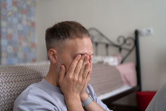 Emotional burnout and exhaustion leave overwhelmed man covering face with hands, sitting alone in bedroom under severe stress and despair.
