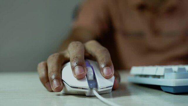Hand using a computer mouse beside a keyboard, Close-up view of a hand using a computer mouse beside a keyboard, with a blurred person in the background and soft moody lighting.