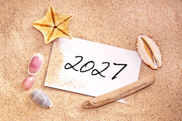 2027 written on a note in the sand with seashells, travel new year card © Delphotostock