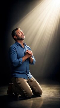 Spiritual awakening and deep faith found in divine beam of light. Kneeling man closes eyes and joins hands in heartfelt prayer, vertical video.