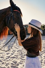 Horse. Horse and man. A woman hugs and touches a black horse in warm backlight on a sandy riding arena  a close-up portrait in profile. © Kateryna