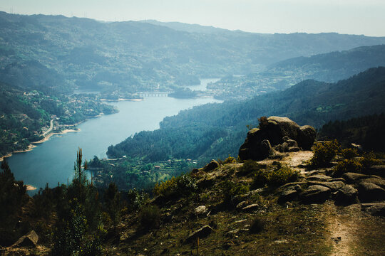 Panoramic view of a mountain reservoir winding through a forested valley with rocky foreground and a distant bridge