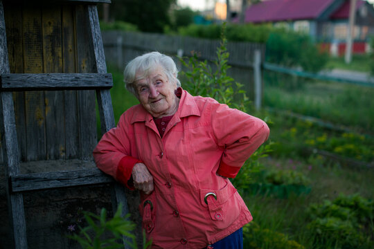 Smiling elderly woman leaning on a weathered wooden ladder beside a rustic house in a garden