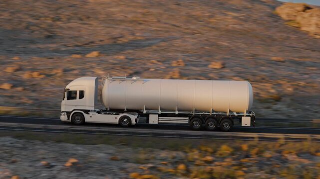 A fixed roadside camera pans to follow a white tanker semi truck rounding a curve and continuing through an expansive landscape of rocky desert hills in warm sunlight.