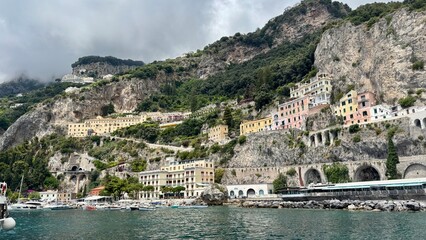 Amalfi harbor on Italy's Amalfi Coast, with colorful buildings climbing steep rocky cliffs, boats moored along the waterfront, a busy promenade and dramatic mountain scenery beneath a cloudy sky © Olshanetskaia