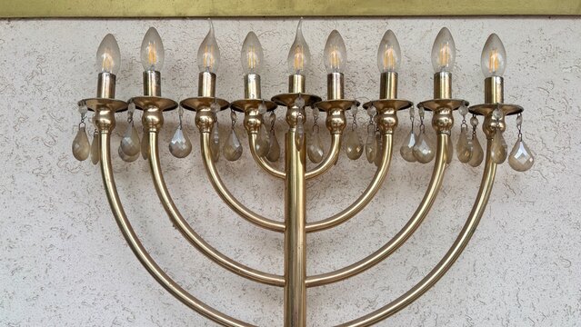 Brass nine-branch hanukkiah menorah with glowing electric candle bulbs, crystal drops and curved arms on a textured white stucco wall, hanukkah, jewish festival of lights and judaism faith concept
