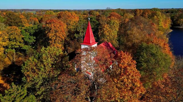 Aerial view of the historic church with a distinctive red roof nestled among vibrant autumn trees near the Daugava river in Lielvarde, Ogre Municipality, Latvia.
