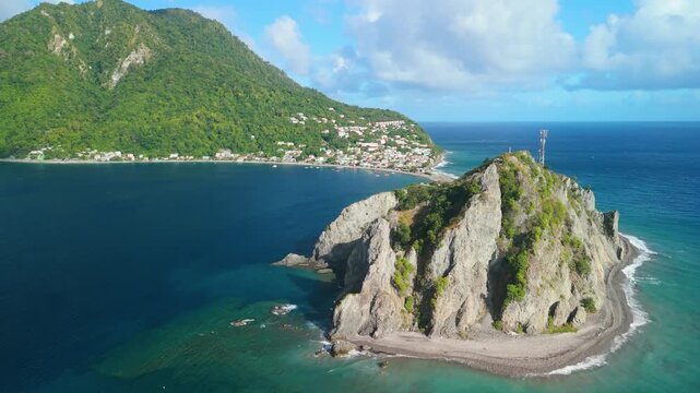 Aerial parallax view as camera moves sideways around rocky Scotts Head headland with communications tower, deep blue Caribbean sea and village behind in Dominica